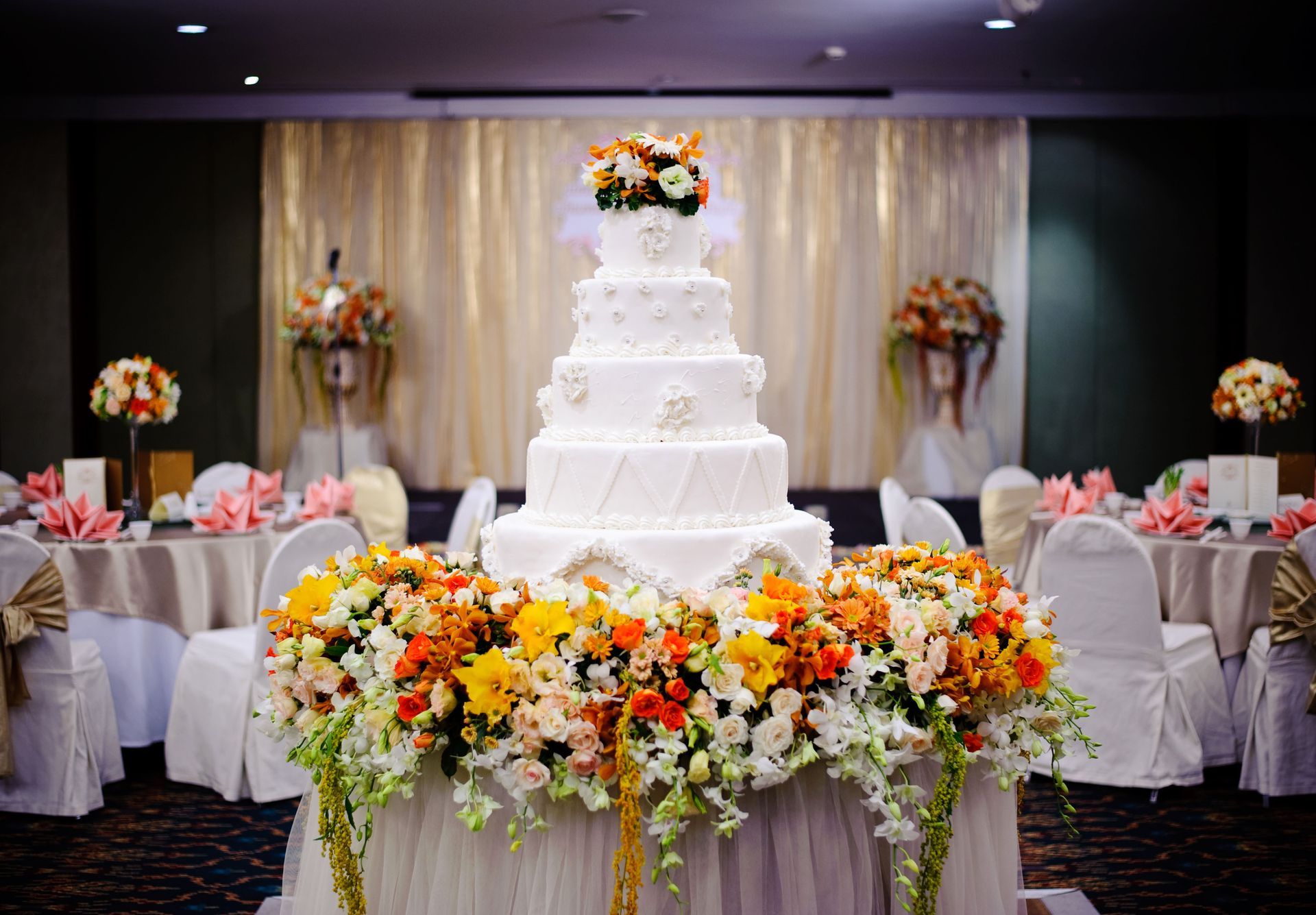 White tiered wedding cake surrounded by colorful flowers in a decorated banquet hall