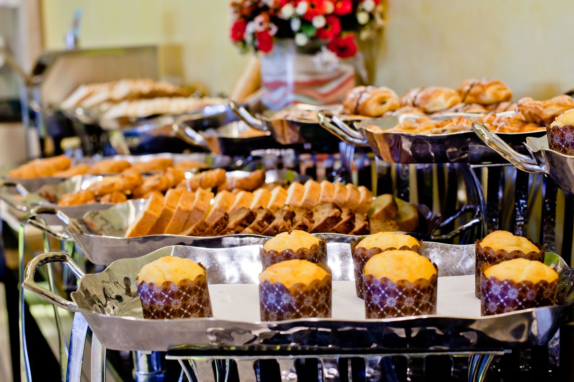 Rows of muffins and pastries on trays at a buffet table, with a floral centerpiece in the background