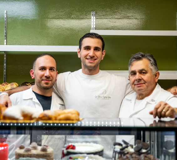 Three bakery staff in white uniforms standing behind a pastry display and smiling.