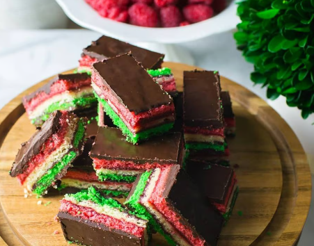 Stack of colorful layered dessert bars with chocolate topping on a wooden board, with raspberries in the background