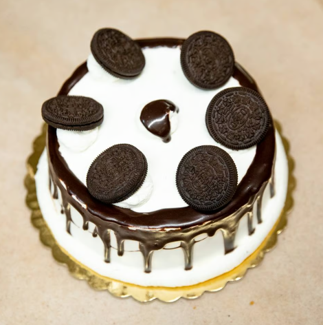 White frosted cake with chocolate drip and Oreo cookies on top, on a gold cake board