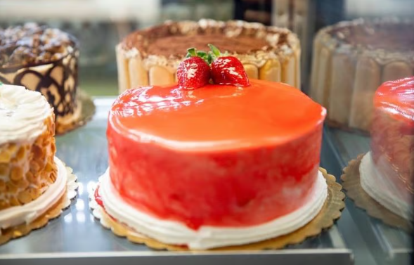 Assorted glossy cakes in a bakery display, with a bright red strawberry-topped cake in front