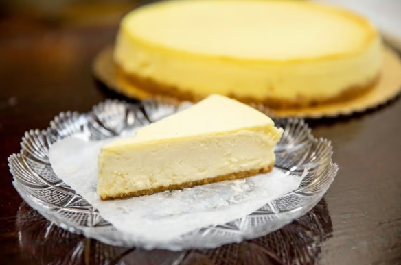Slice of cheesecake on a glass plate with parchment, with a whole cheesecake blurred in the background