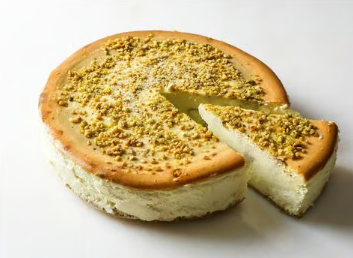Round pistachio cake with a slice removed, topped with chopped nuts on a white background