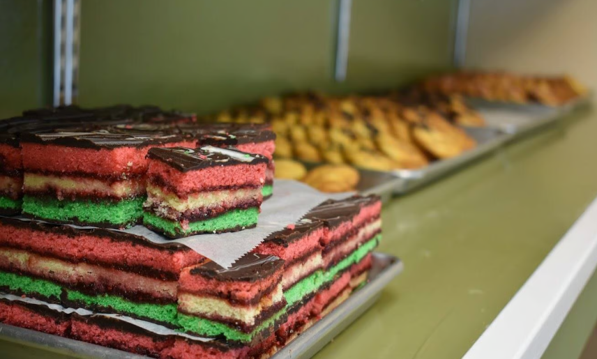 Stacked colorful layered cakes on a kitchen counter with trays of baked goods in the background
