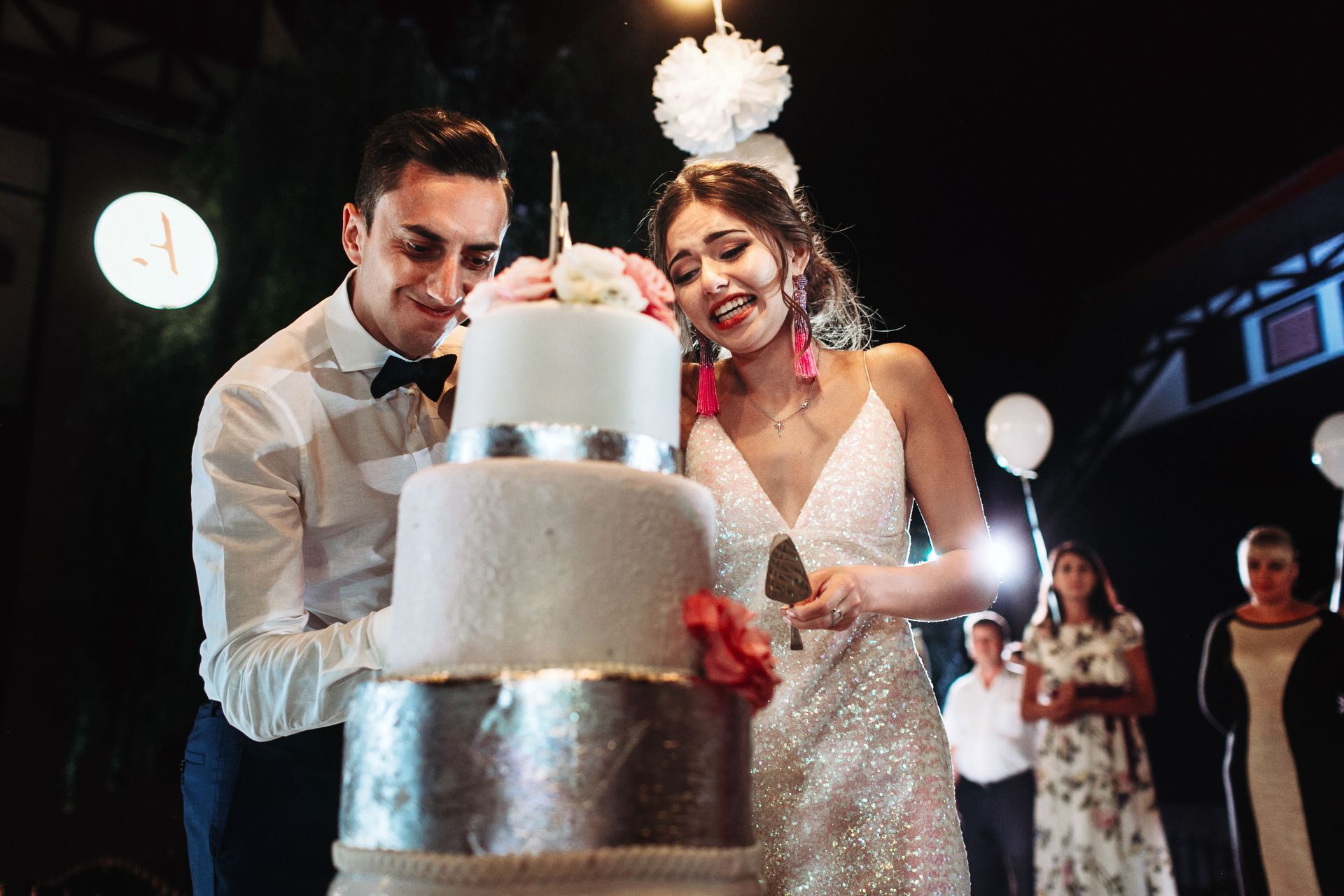 Couple cutting a white wedding cake at a celebration, with guests and balloons in the background