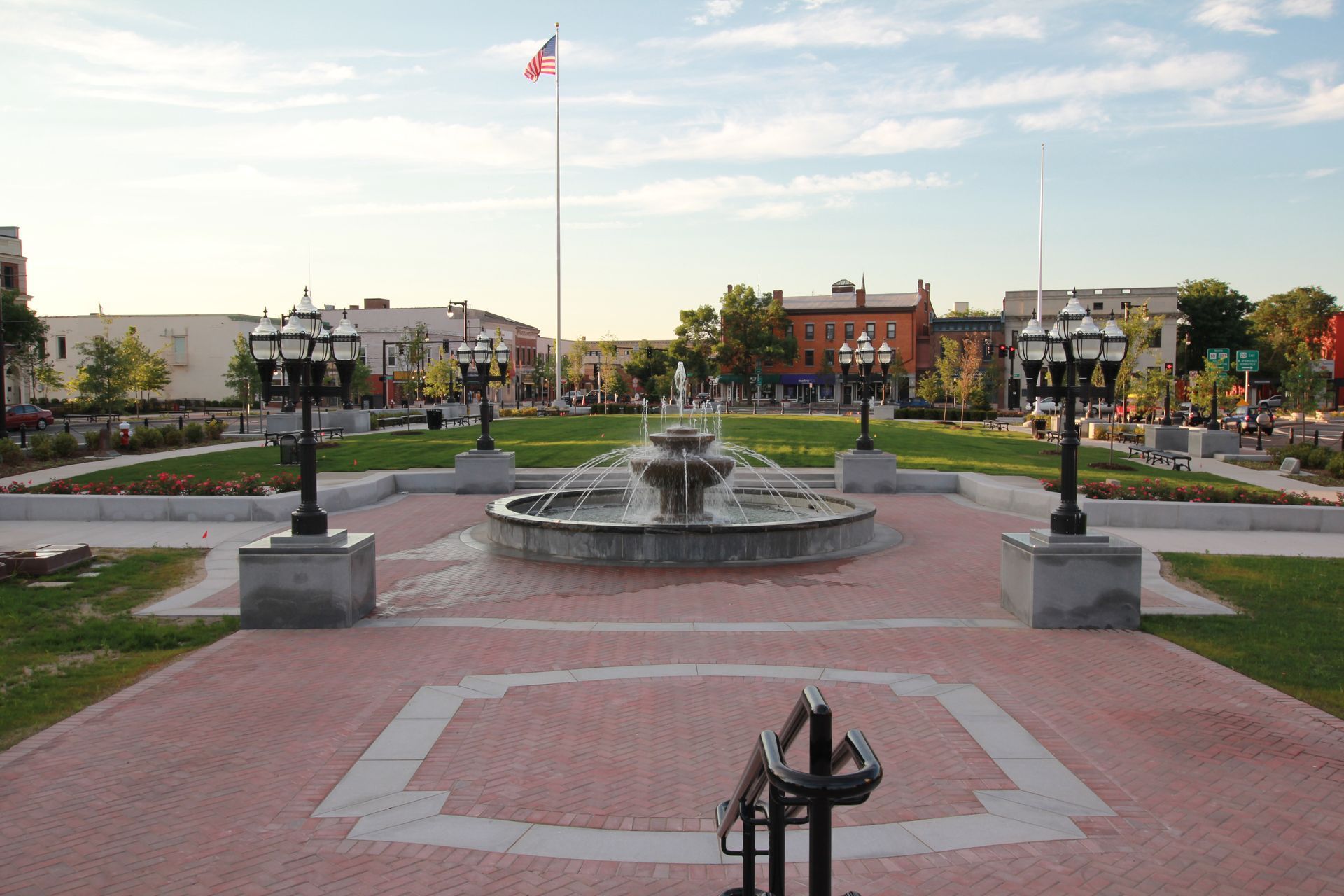 A brick plaza with a fountain, surrounded by flower beds, lampposts, and buildings. Flag flies in the distance.