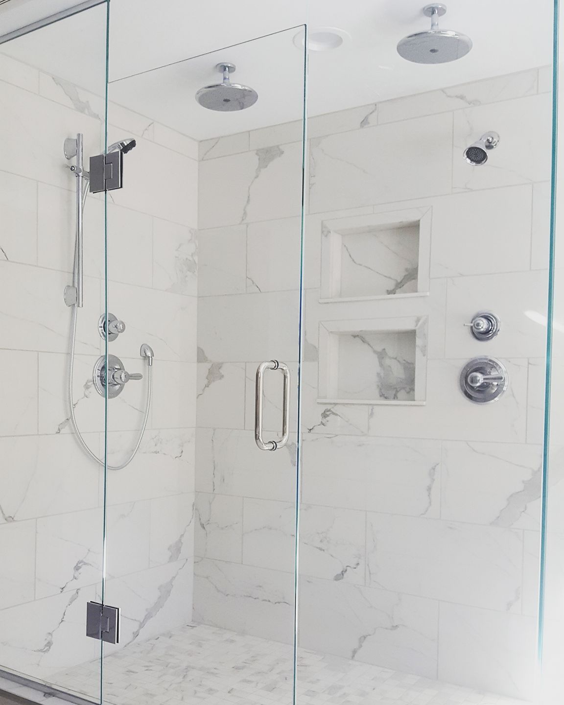 Shower with white marble tiles, glass doors, and two overhead showerheads.