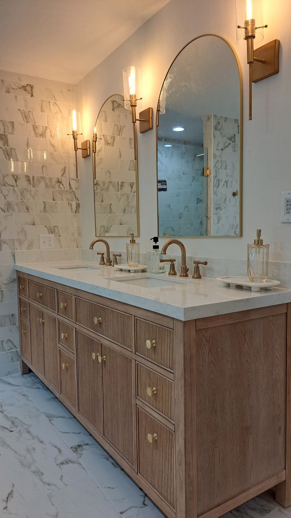 Bathroom with a double vanity, gold fixtures, arched mirrors, and marble tile.