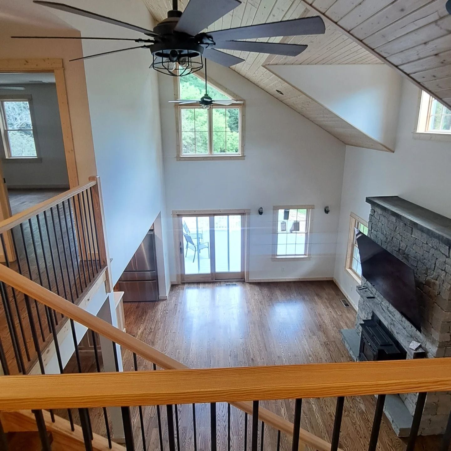 Interior view of a living room with wooden floors, fireplace, and tall windows. A staircase is in the foreground.