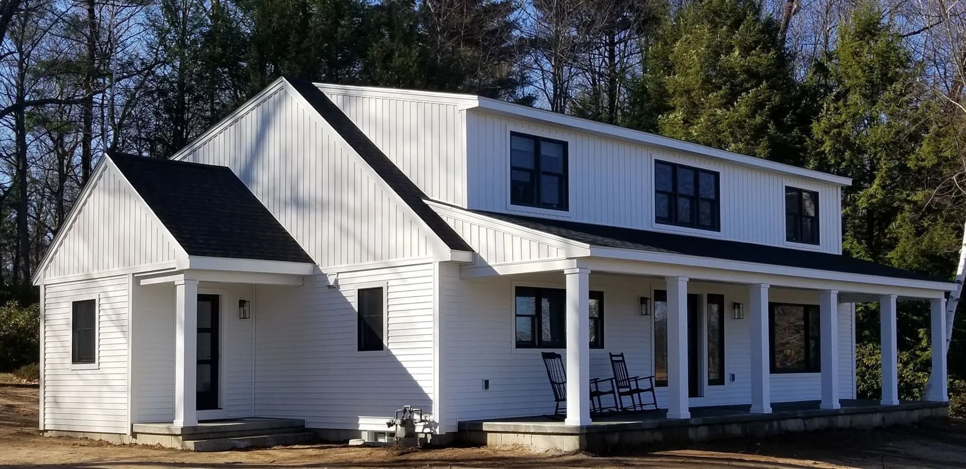 White two-story house with black trim and a wraparound porch, set in front of trees.