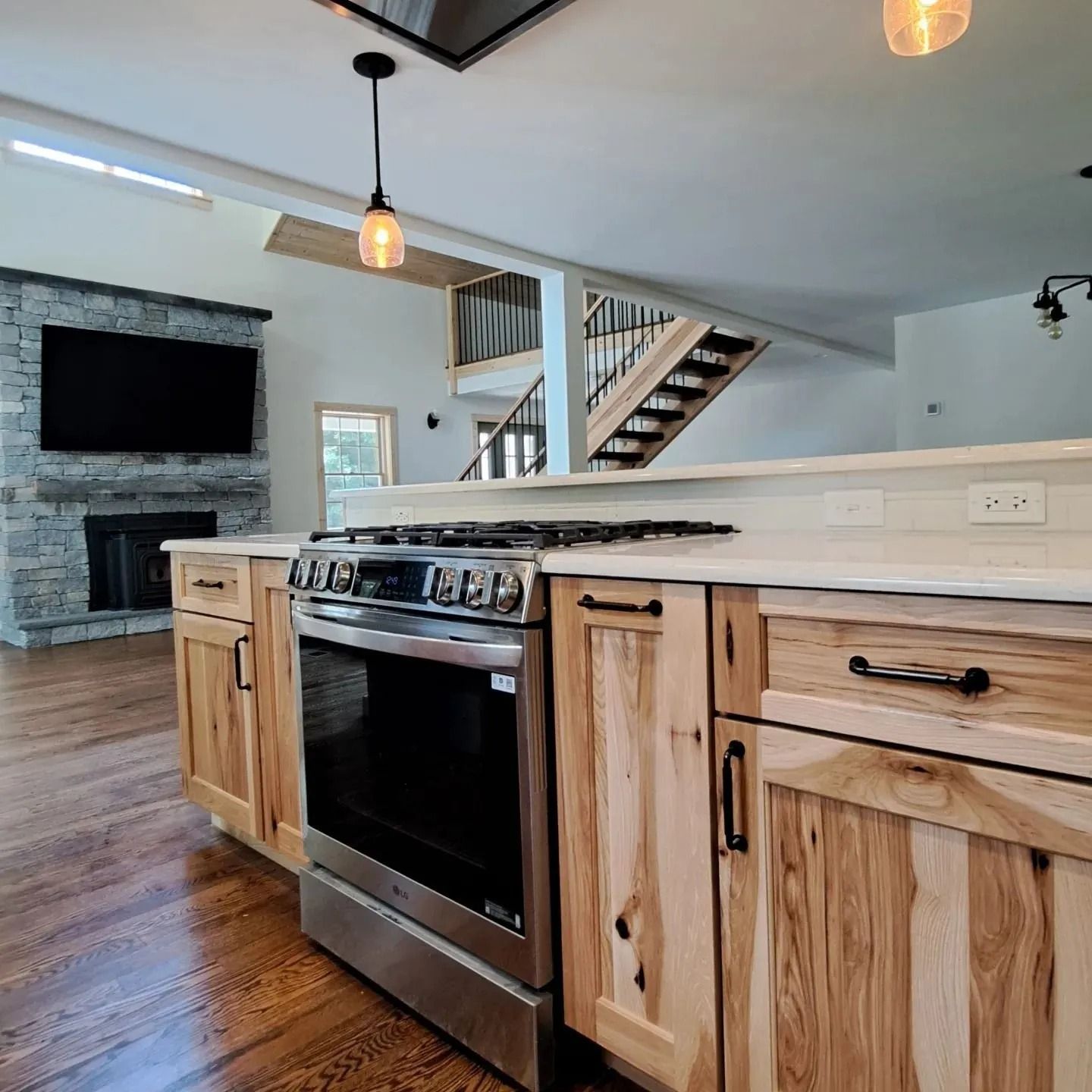 Kitchen island with stove and light wood cabinets, fireplace, and staircase in the background.