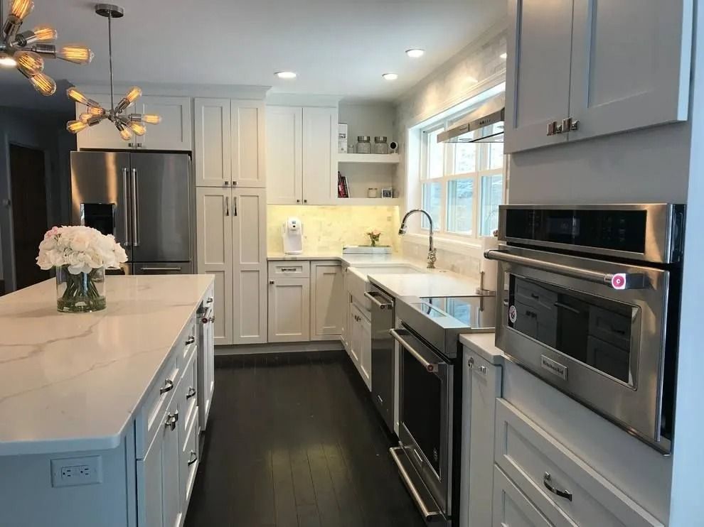 Modern white kitchen with island, stainless steel appliances, dark wood floor, and overhead lighting.