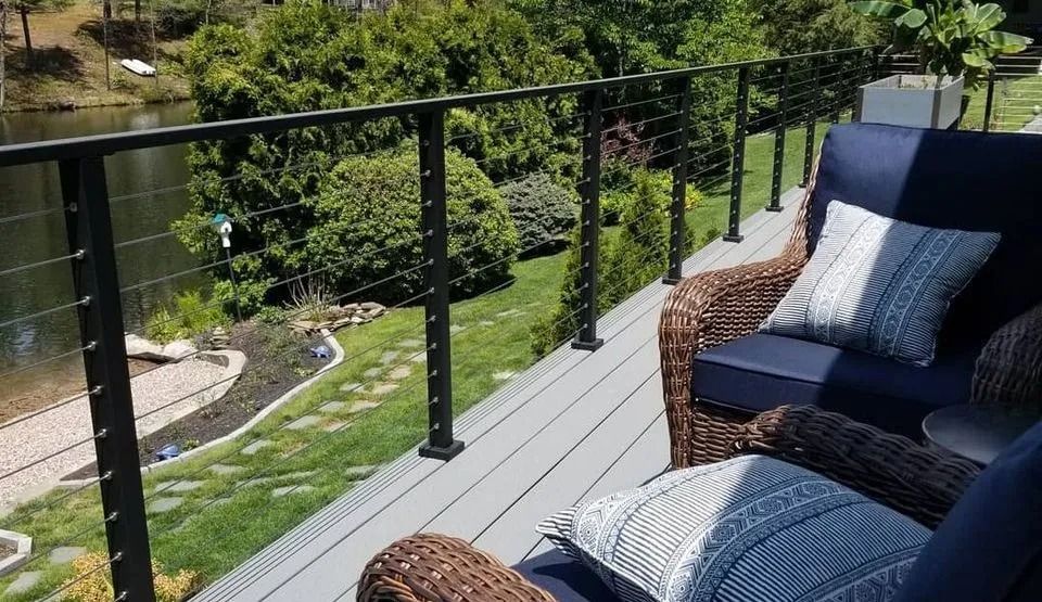 A deck with black cable railing and two blue cushioned wicker chairs overlooking a lake and greenery.