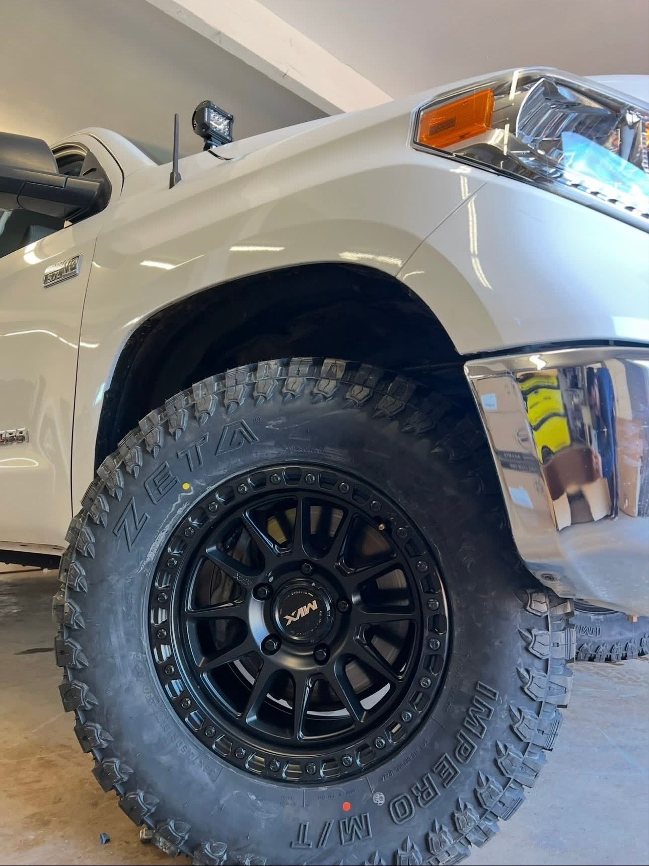 Close-up of a white truck’s front wheel with black off-road tire and rim in a garage.