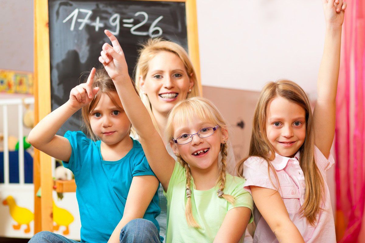 Three children sitting in a classroom, smiling in front of a chalkboard with a math problem.