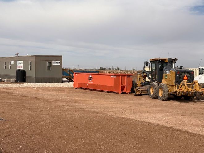 A metal dumpster filled with construction debris, situated at a residential building site under a clear blue sky.