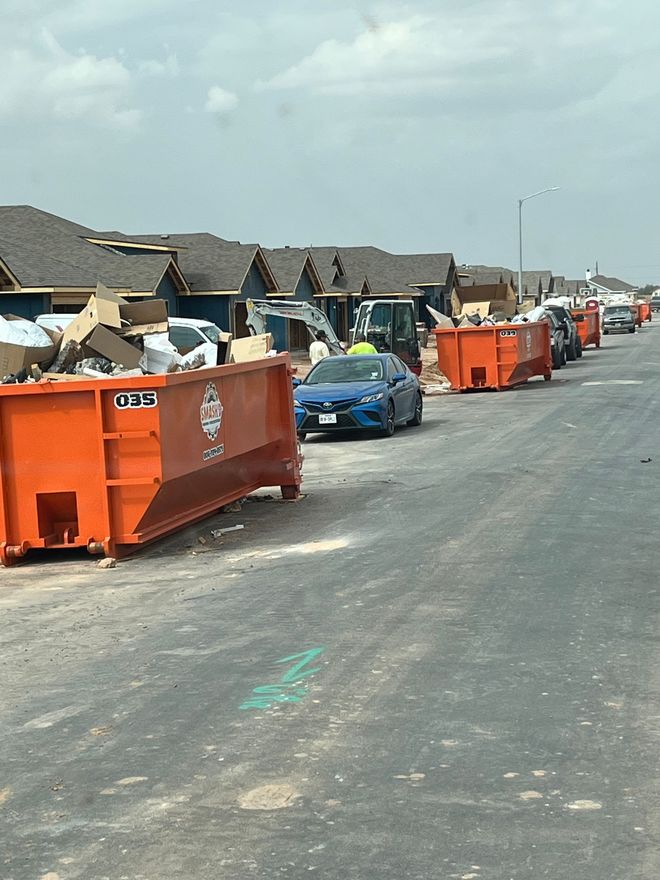 A large, open-top metal dumpster filled with wooden debris and trash, situated outdoors with a work truck in the background.