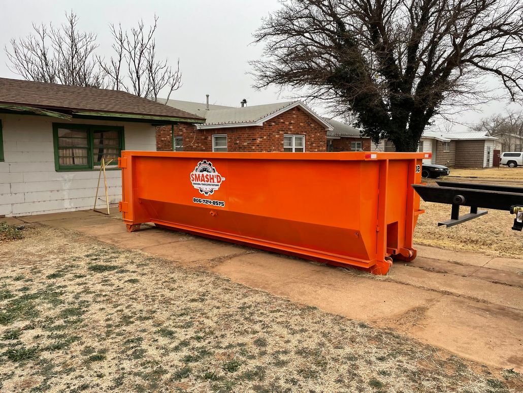 An empty, bright orange roll-off dumpster sitting on a gravel surface next to a white metal building.
