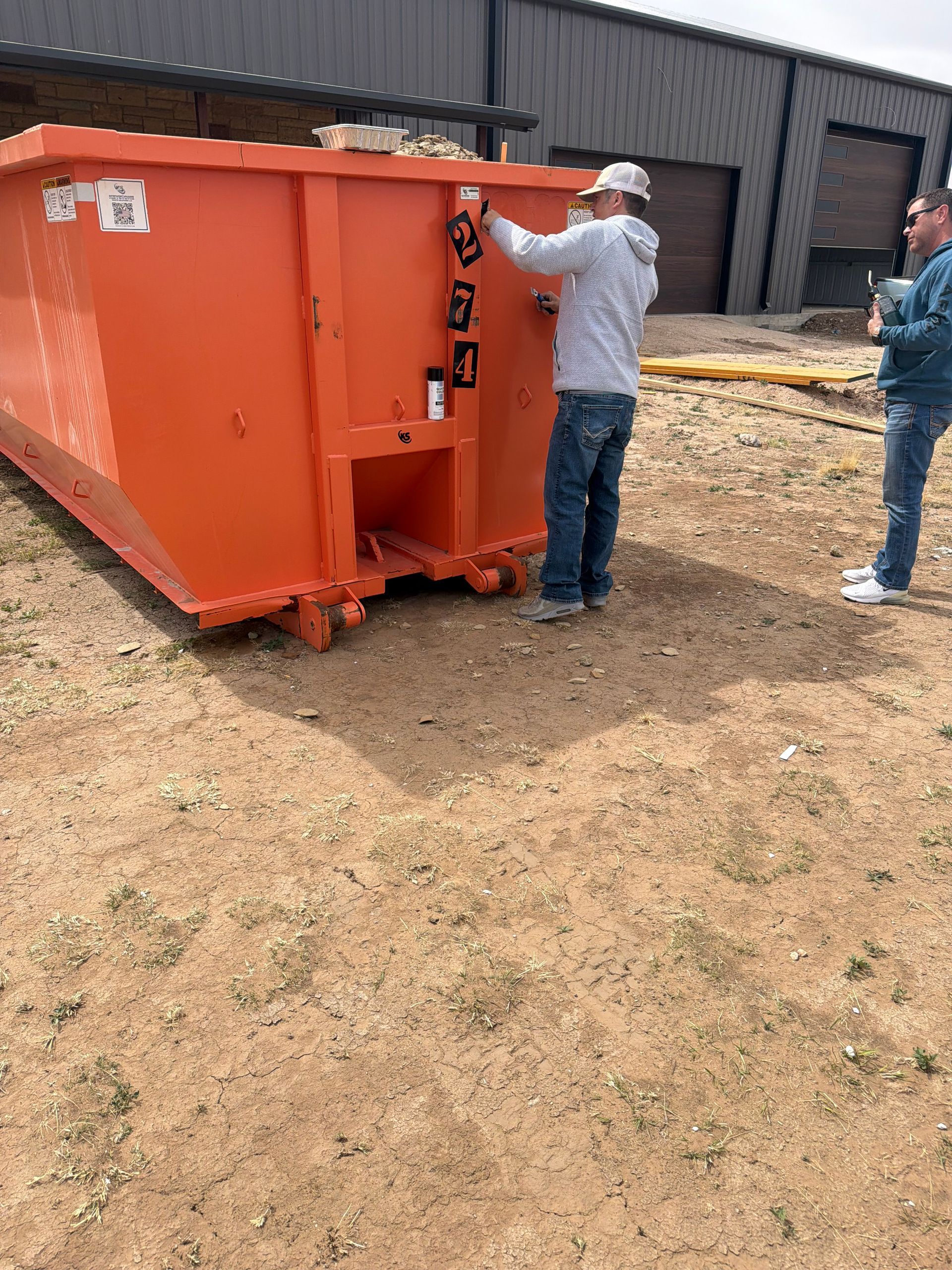 Two people paint numbers on an orange metal dumpster parked on a dirt lot outside a warehouse.