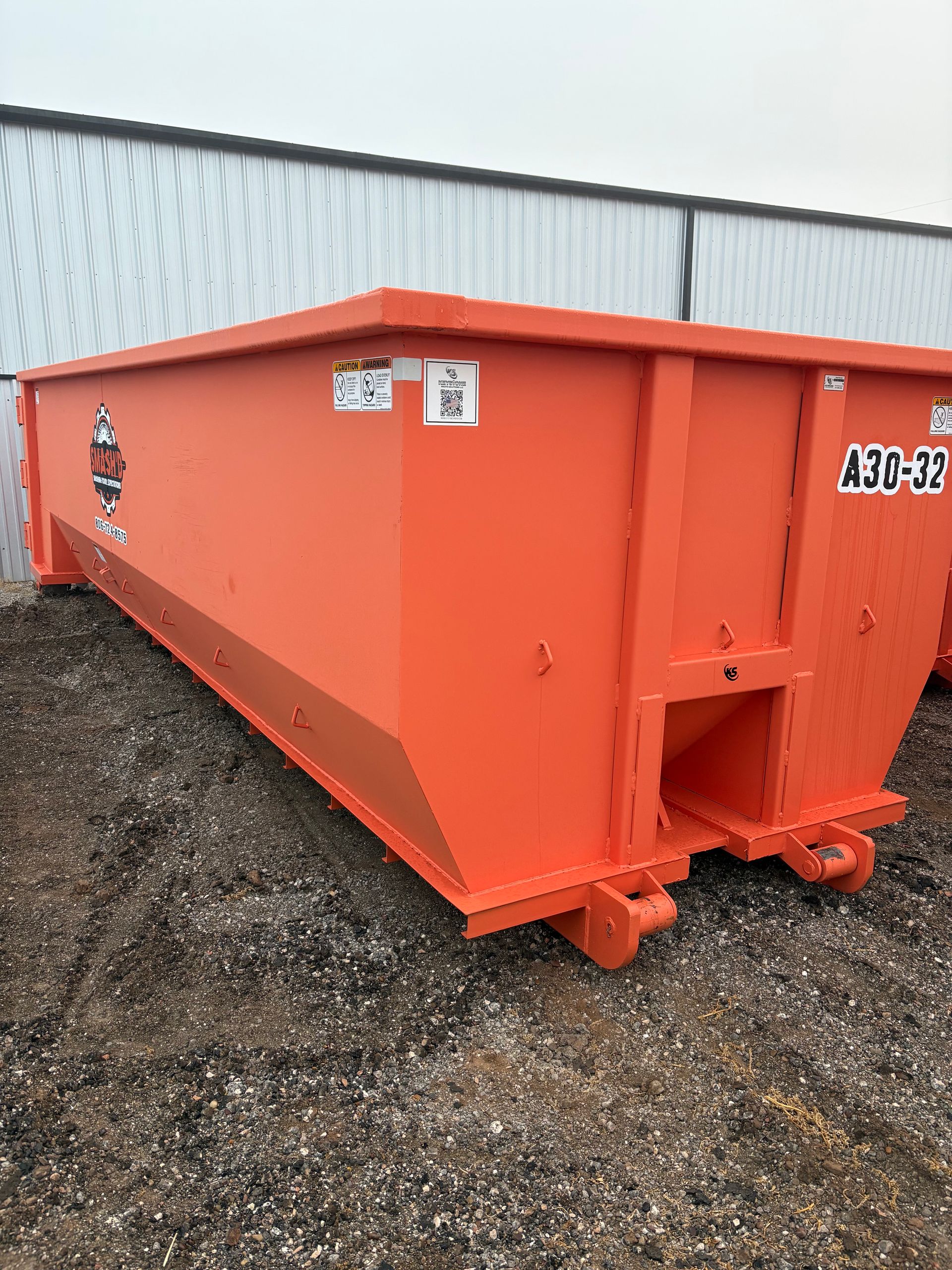 A bright orange, rectangular roll-off dumpster sitting on a gravel lot in front of a white metal building.