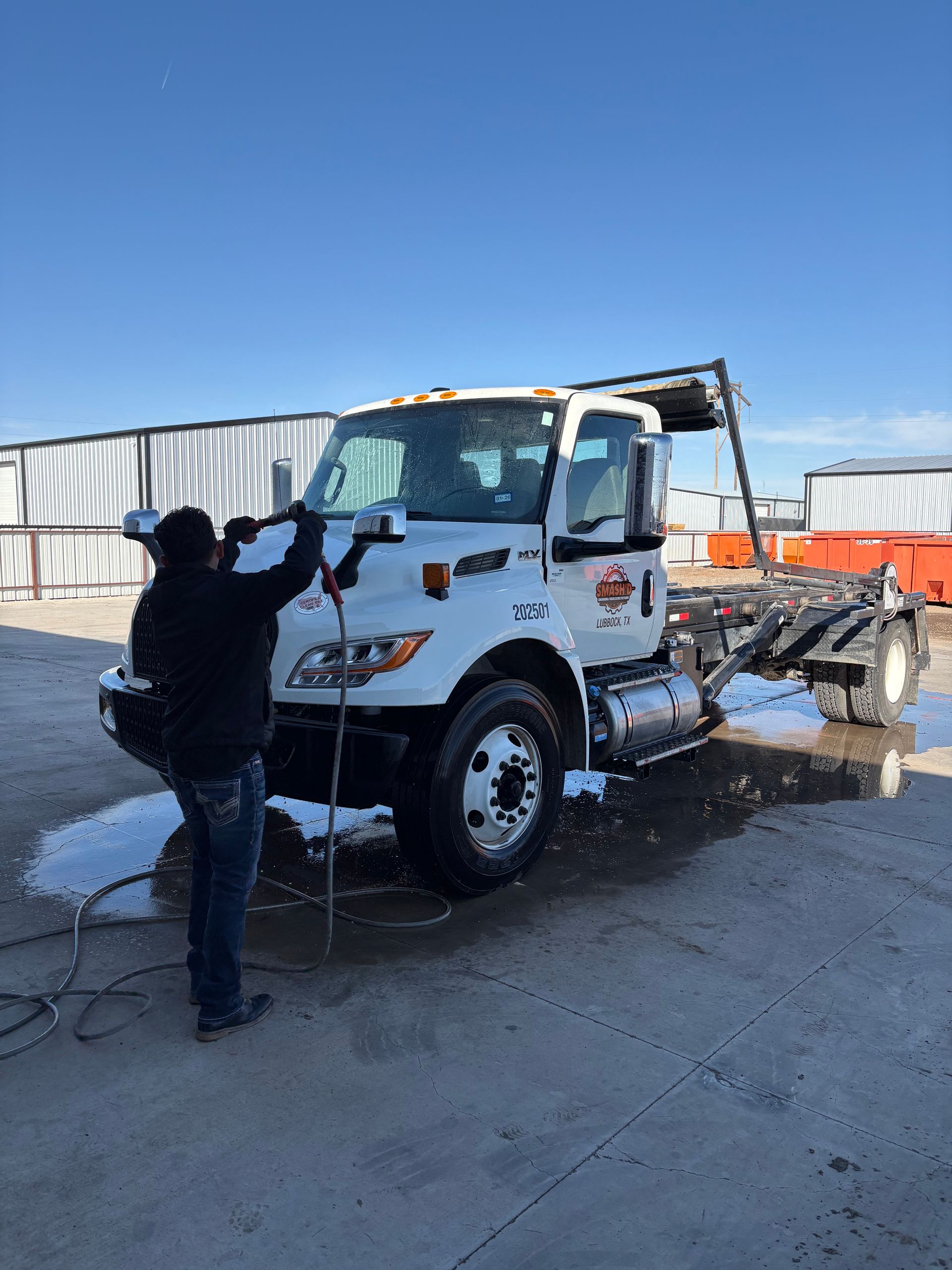 A worker uses a pressure washer to clean a white commercial truck parked on a concrete lot under a clear blue sky.