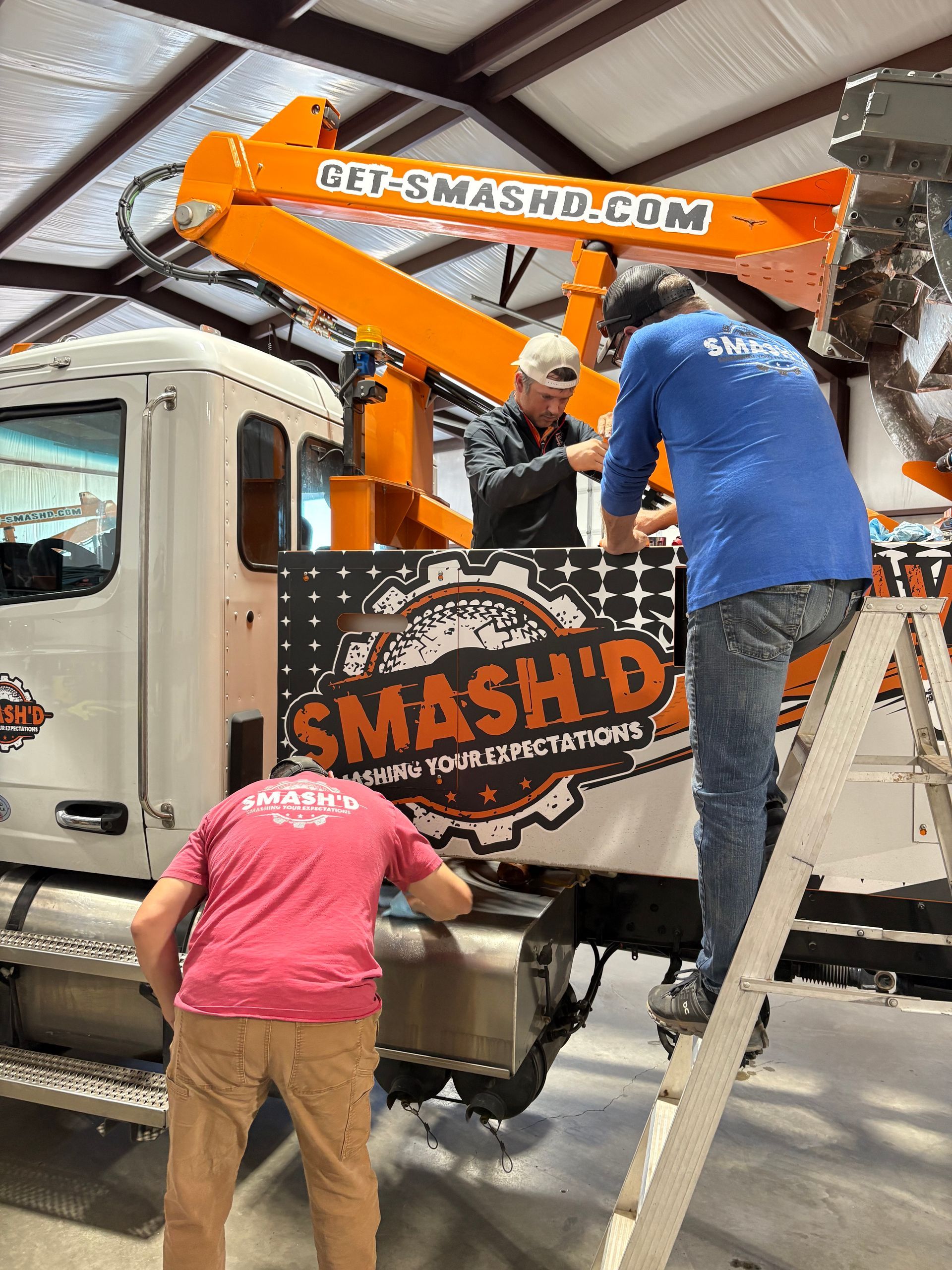 Three people working on a large white truck with a bright orange tow boom in an indoor garage.