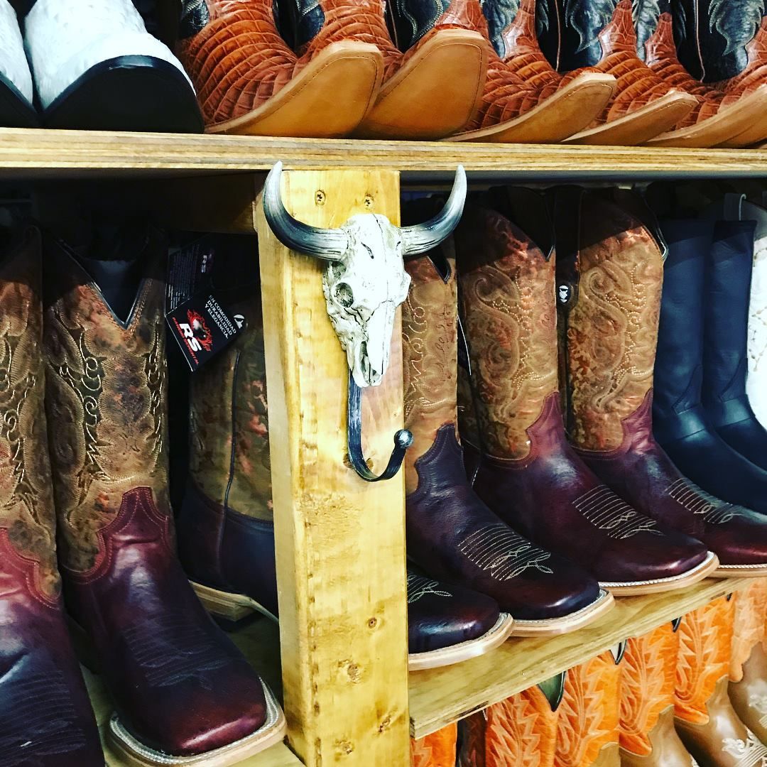 A row of cowboy boots are lined up on a shelf with a bull skull hanging on a wooden post.