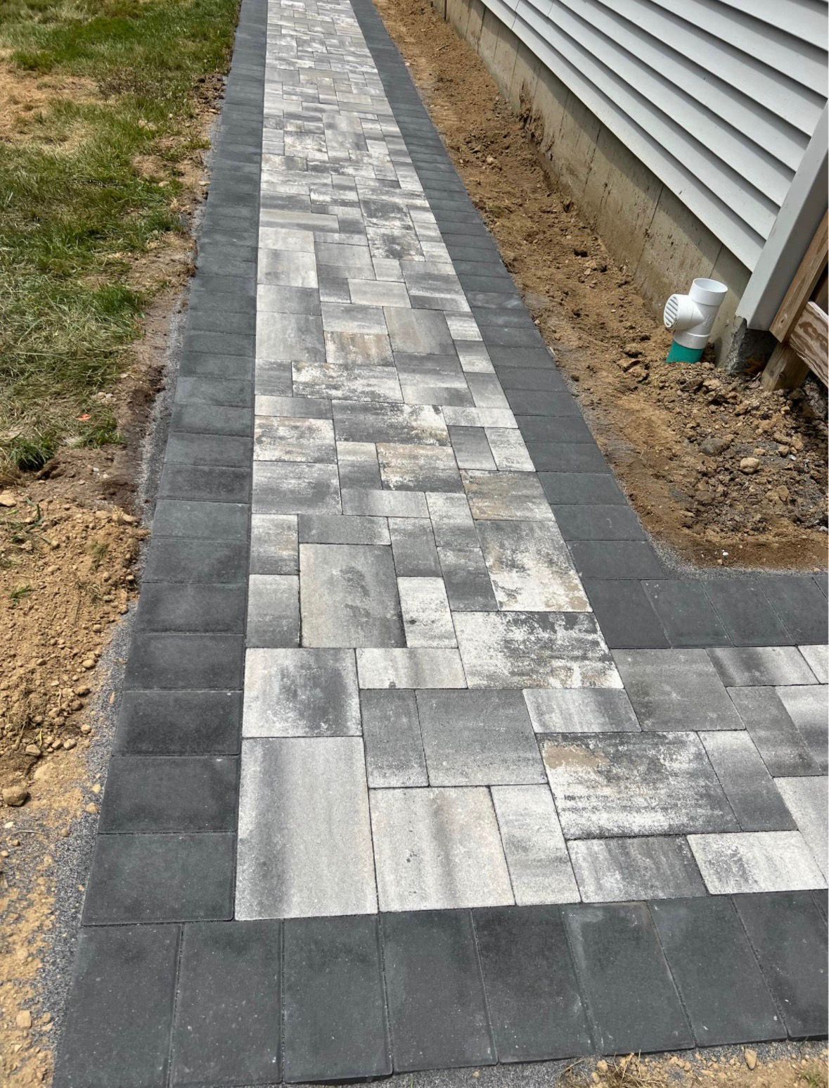 Gray paver walkway beside a house, bordered by dark stones and unfinished soil.