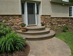 Curved stone front steps leading to a gray door with potted plants and landscaped garden beds