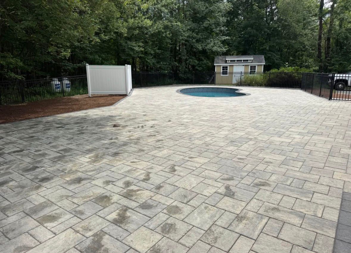 Stone patio with a round blue pool and white shed, bordered by trees and a black fence