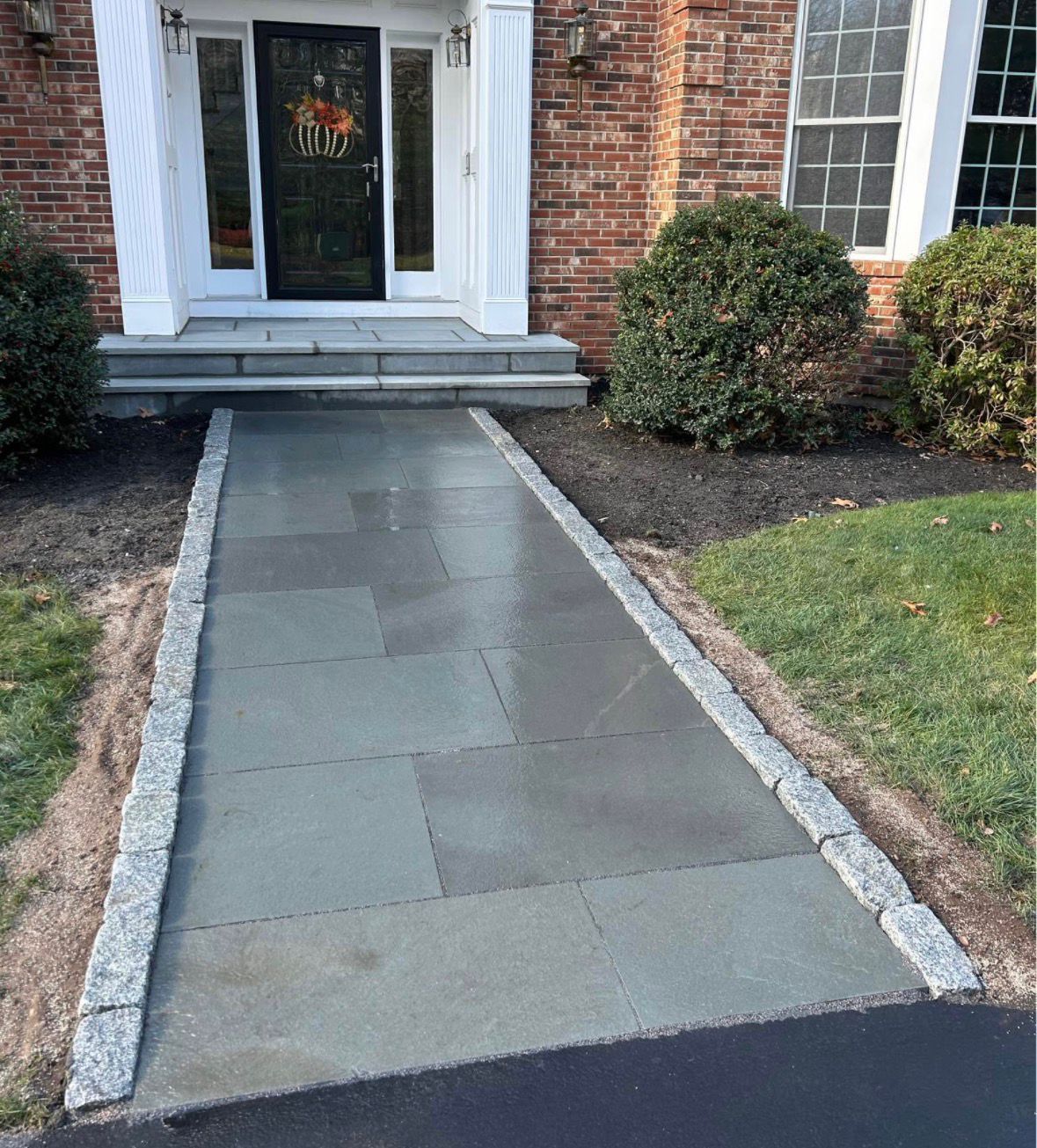 Front walkway with concrete path leading to a brick house entrance, bordered by stone edging and lawn