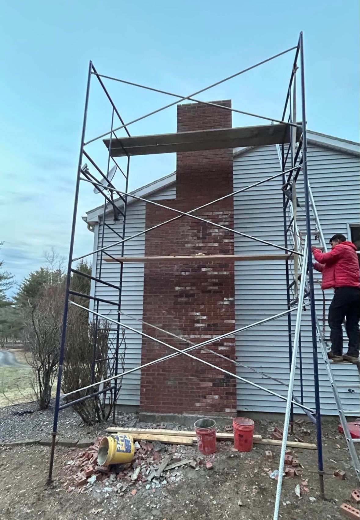 Scaffolding around a brick chimney on a house exterior, with a worker on the right.