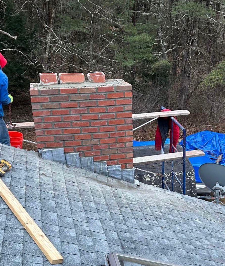 Roof under construction with brick chimney, workers, and blue tarp in a wooded area
