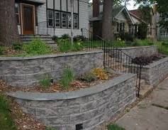 Curved stone retaining wall with garden beds and a walkway beside a house and sidewalk