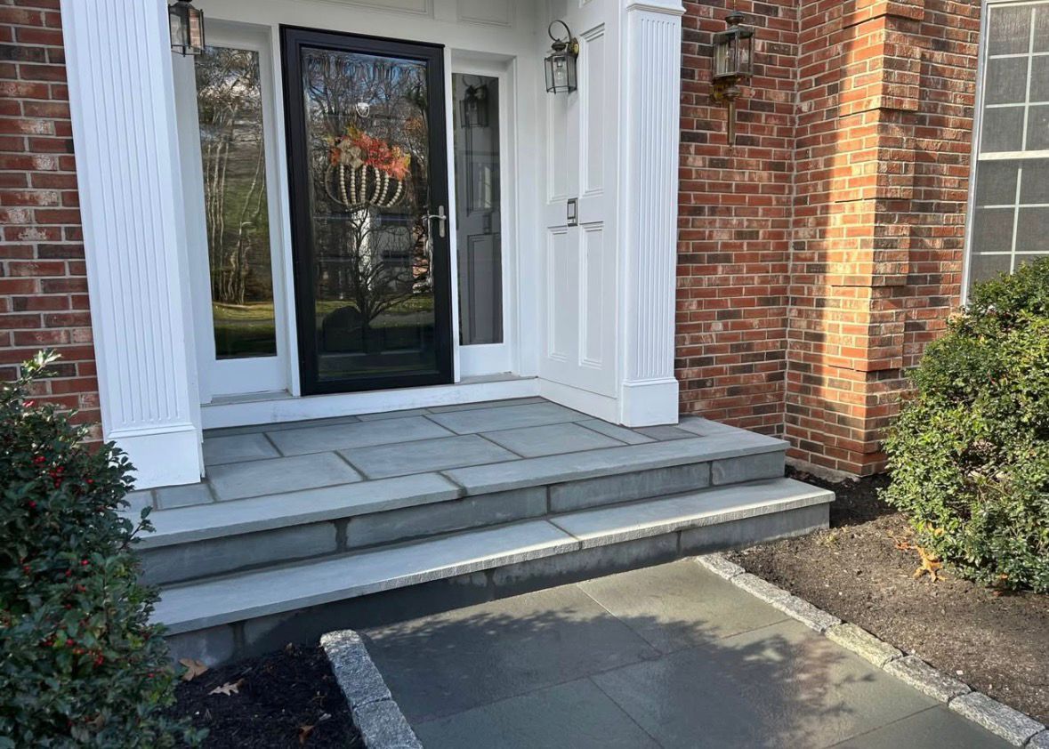 Front porch with gray steps, black door, white columns, and brick exterior beside a driveway.