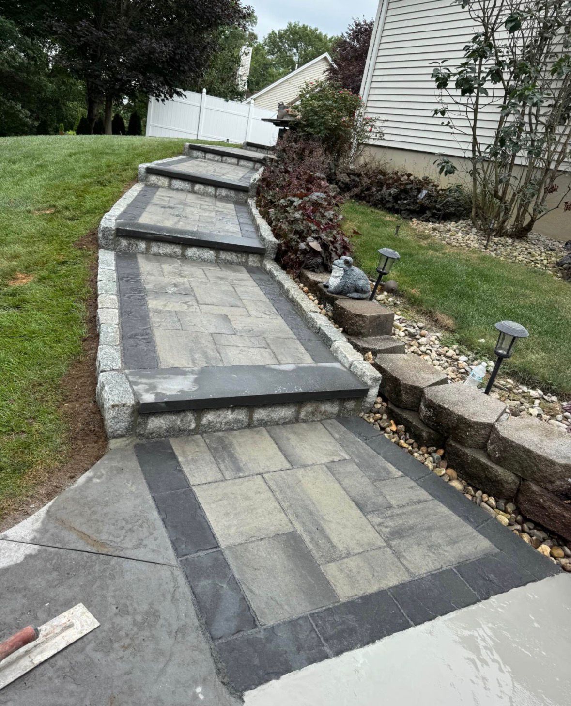 Stone walkway with steps and a dark border, running beside a house and landscaped yard.