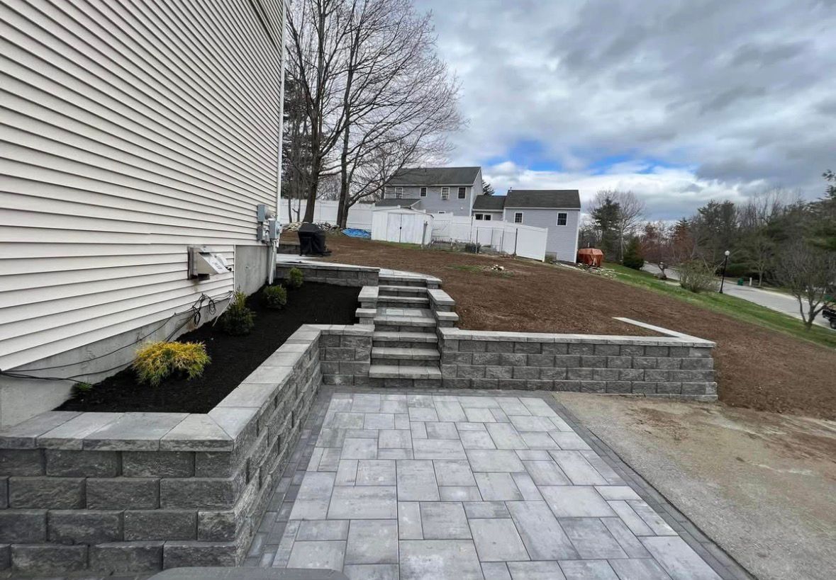 Stone patio and steps beside a white house, leading to a dirt driveway and suburban homes.