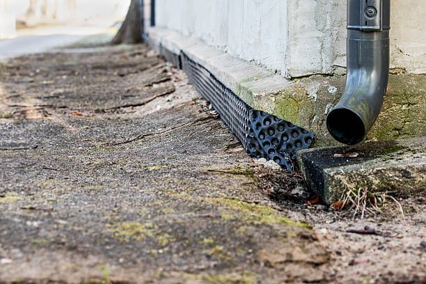 Drainpipe beside a white wall above a black perforated drainage strip on a gravel path