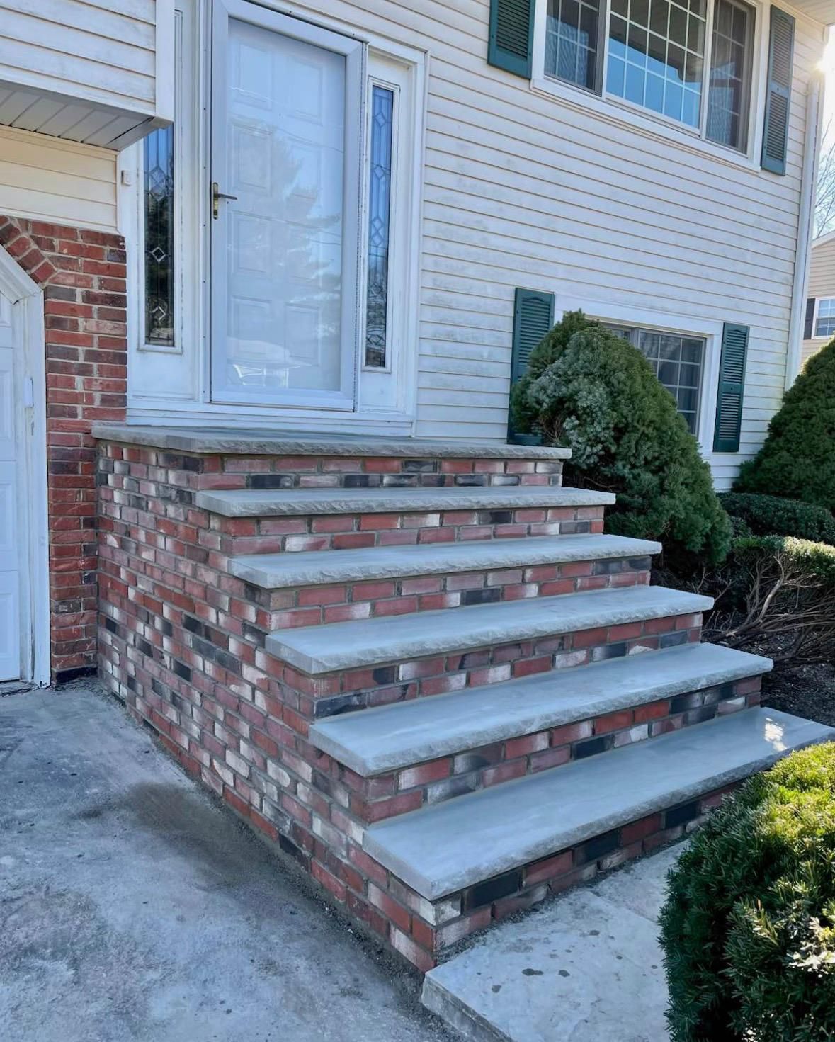Brick front steps leading to a white house entry with a glass storm door and shrubs nearby