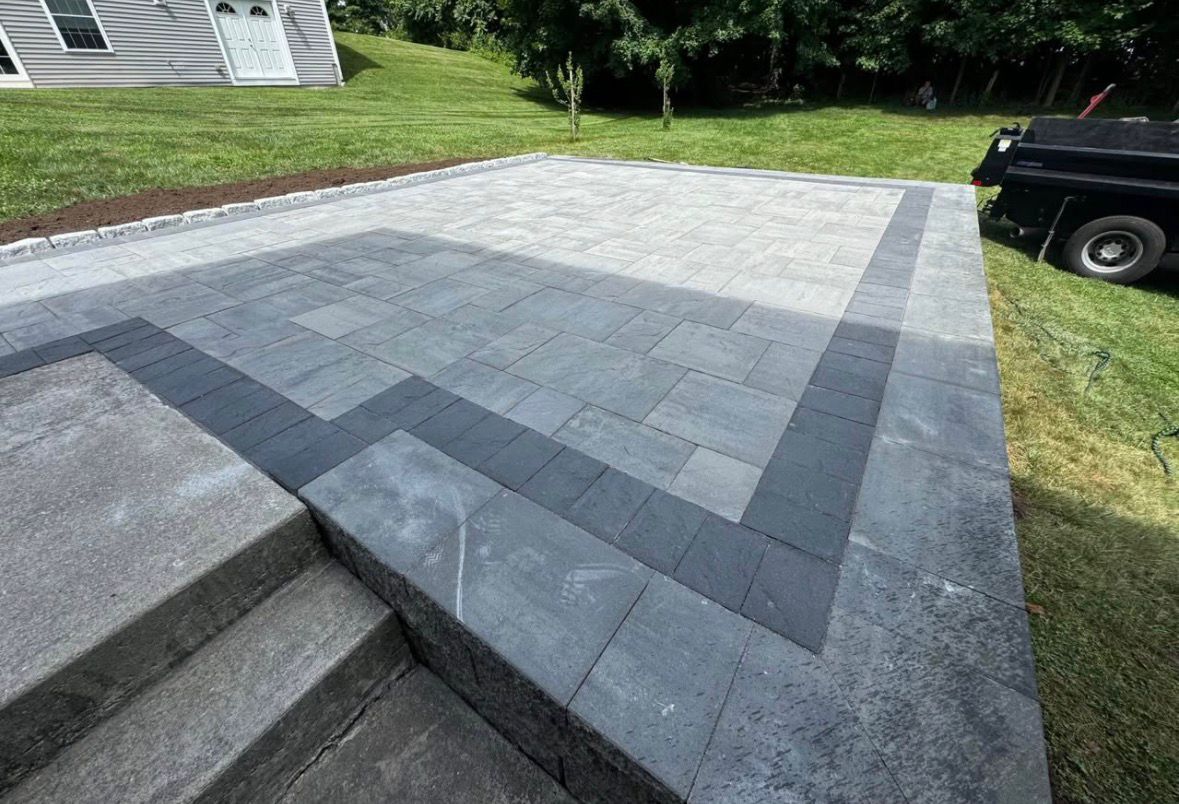 Gray paver patio with dark border beside house steps and lawn, next to a black truck