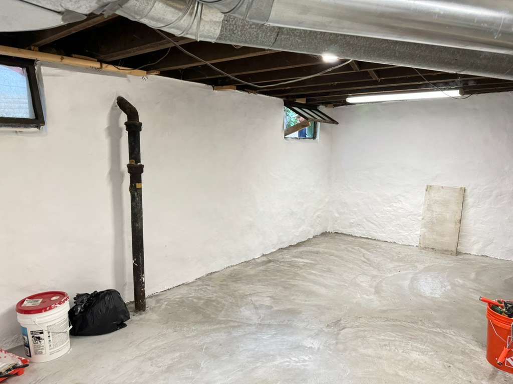 Empty basement with white walls, concrete floor, exposed ceiling beams, and a black pipe near a corner.