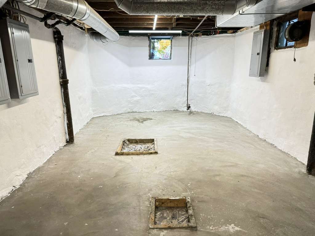 Empty basement with white walls, exposed ceiling beams, and two square floor openings near the center.
