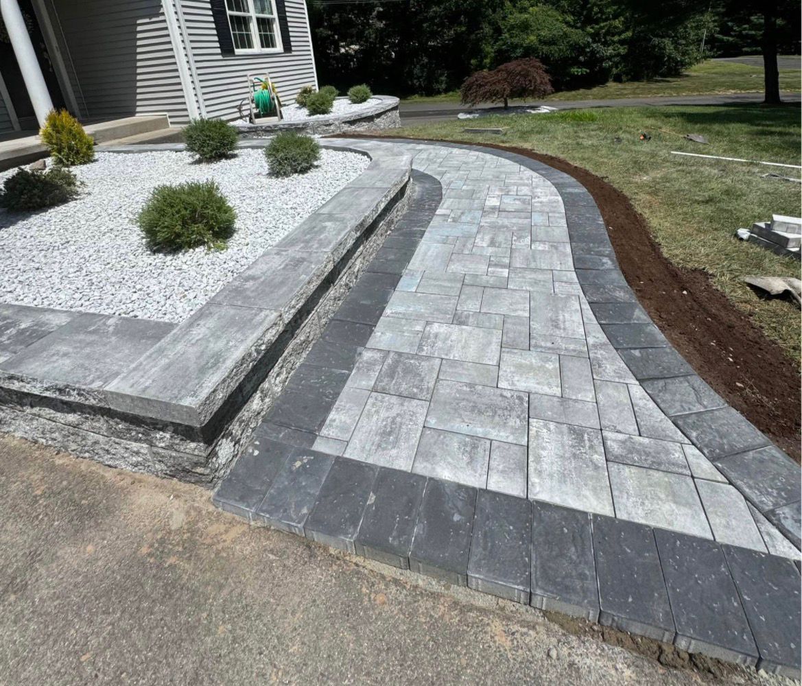 Curved gray paver walkway beside a house, bordered by white gravel and mulch in a landscaped yard