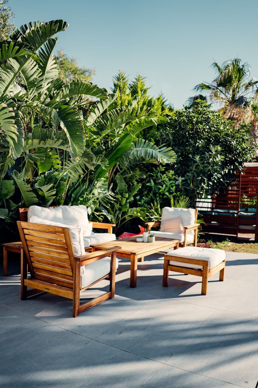Wooden patio chairs and ottoman on a sunny terrace surrounded by lush tropical plants