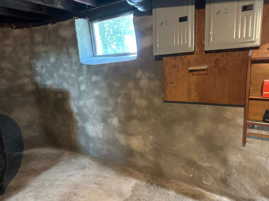 Basement wall with window, utility panels, and brown cabinets under exposed ceiling beams.