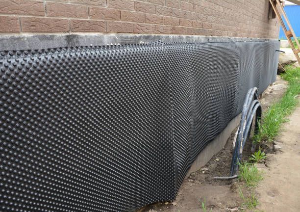 Black dimpled waterproofing membrane along a brick foundation wall beside a narrow dirt trench.