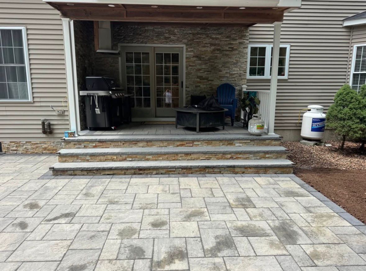 Covered backyard patio with stone steps, paver walkway, and outdoor furniture against a house exterior