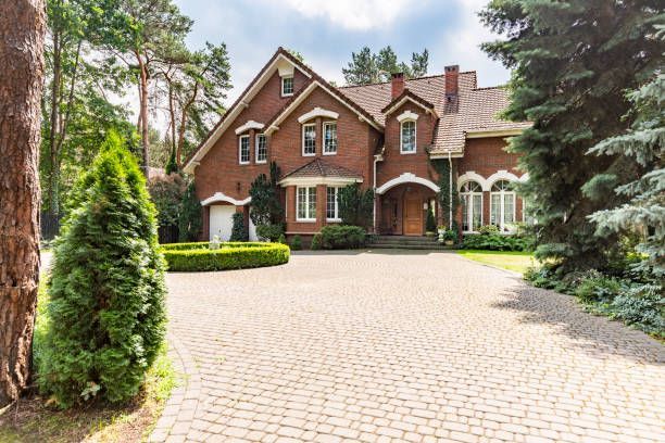Brick house with arched windows and a large paved driveway, framed by trees and greenery.