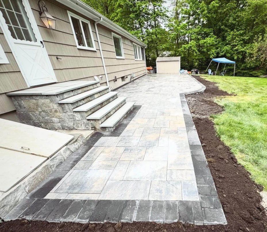Freshly paved stone walkway beside a house with front steps and a lawn under trees